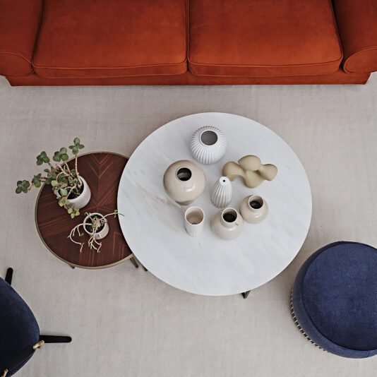 Top-view of a contemporary living room showcasing the Modern Set Of Round Coffee Tables adorned with beige vases and a small potted plant. Nearby, an additional smaller round table holds another potted plant. The room is furnished with a red sofa and two dark blue chairs.