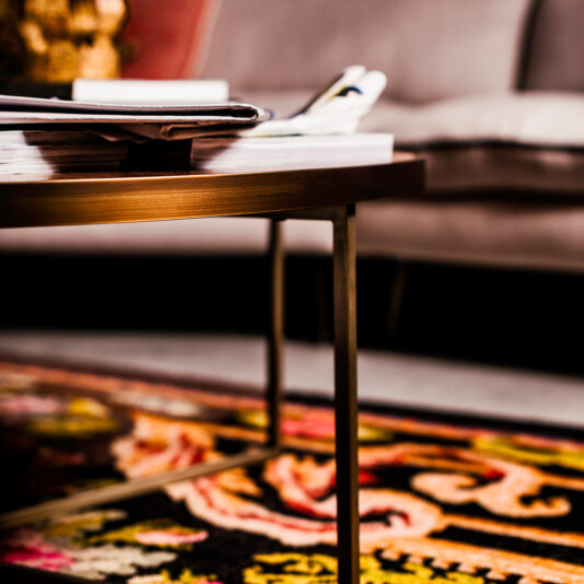 A close-up view of the Round Walnut Veneer Coffee Table Set with golden legs, partially covered with magazines and books. The table stands on a vibrant, patterned rug, with a blurred gray sofa visible in the background. The setting appears to be a cozy living room.