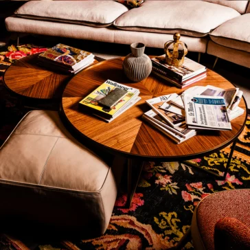 A cozy living room features a colorful rug, the Round Walnut Veneer Coffee Table Set with books, magazines, and a decorative vase on top. Nearby are a beige ottoman and a plush-cushioned sofa creating a warm and inviting atmosphere.