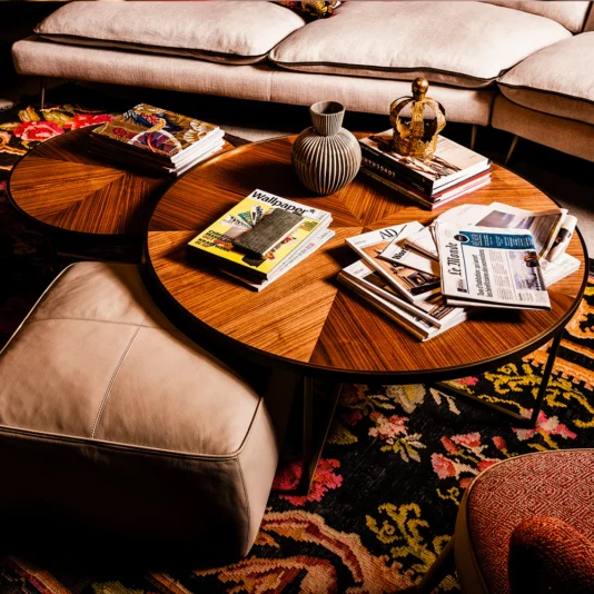 A cozy living room features a colorful rug, the Round Walnut Veneer Coffee Table Set with books, magazines, and a decorative vase on top. Nearby are a beige ottoman and a plush-cushioned sofa creating a warm and inviting atmosphere.
