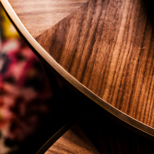 Close-up of the Round Walnut Veneer Coffee Table Set, showcasing its rich brown wood grain enhanced by soft lighting. The table's metallic edge adds a modern touch, while a blurred, colorful object can be seen in the background to the left.