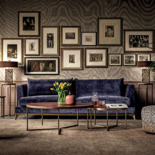 A stylish living room features a navy blue velvet sofa and the Round Walnut Veneer Coffee Table Set, which holds a vase of yellow tulips and drinks. The backdrop includes a wall adorned with framed art, adding a sophisticated touch. Two lamps flank the sofa, illuminating the cozy space.