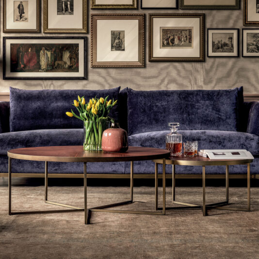 A cozy living room with two velvet blue sofas, a Round Walnut Veneer Coffee Table Set, and a matching side table. The coffee table features a vase of yellow flowers and a pink teapot, while the side table holds a decanter, two glasses, and a book. Framed art decorates the wall.