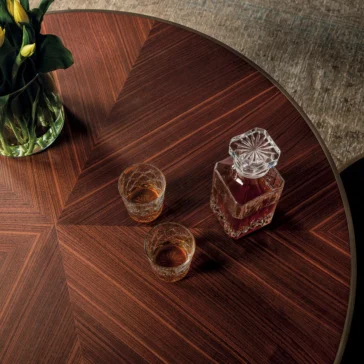 The Round Walnut Veneer Coffee Table Set features a starburst pattern, holding a crystal decanter with dark liquid and two glasses. A vase of yellow calla lilies decorates one side, gently illuminated to enhance the rich wood grain.