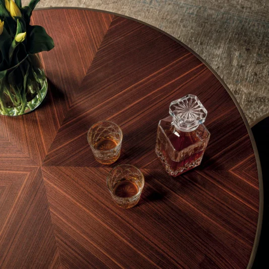 The Round Walnut Veneer Coffee Table Set features a starburst pattern, holding a crystal decanter with dark liquid and two glasses. A vase of yellow calla lilies decorates one side, gently illuminated to enhance the rich wood grain.