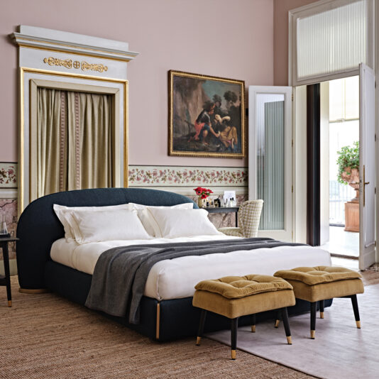 Small Button Upholstered Velvet Bench A stylish bedroom featuring a large bed with white bedding and a dark upholstered headboard. The Small Button Upholstered Velvet Bench sits gracefully at the foot of the bed. The room boasts ornate wall molding, a painting, a desk with a chair, and an open door leading to a balcony filled with natural light.