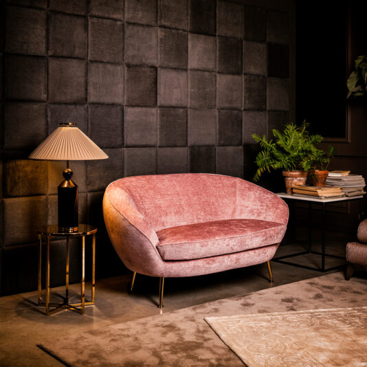 Small Contemporary Designer Sofa A Small Contemporary Designer Sofa in pink velvet with gold legs anchors the modern living room, set against a black textured wall. To the left, a round side table with a lamp sits on a beige rug. On the right, a black table holds a potted plant and a stack of books.