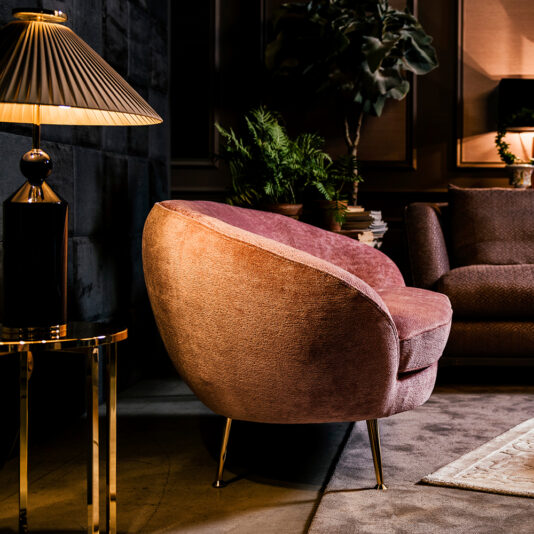Small Contemporary Designer Sofa A cozy, modern living room with a plush, round pink armchair on sleek metal legs. The room features warm lighting from a table lamp with a pleated lampshade, a leafy potted plant, and the Small Contemporary Designer Sofa in the background, creating an inviting and elegant ambiance.