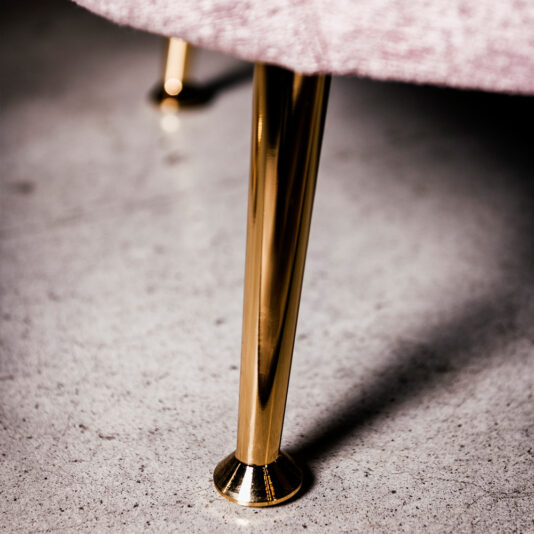Small Contemporary Designer Sofa Close-up image of the gold-colored metal leg of the Small Contemporary Designer Sofa, which is placed on a polished concrete floor. The leg supports fabric that appears to be light pink or mauve. The focus is on the shiny gold leg contrasting with the textured floor.