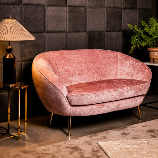 A stylish interior showcases a Small Contemporary Designer Sofa in plush pink velvet with gold legs, set against a dark, textured wall. A round side table with a warm-toned lamp sits to the left, while a green potted plant adds nature's touch in the background.