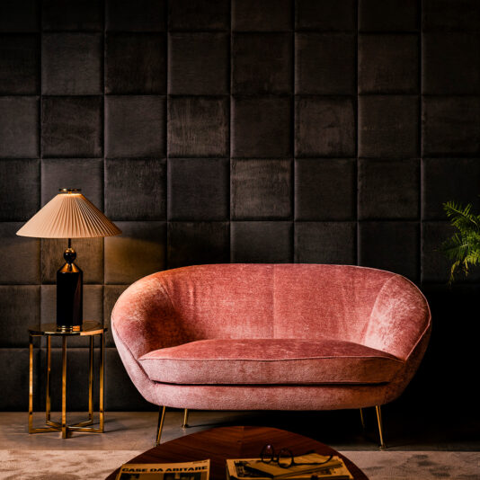 Small Contemporary Designer Sofa A stylish room features a cozy small contemporary designer sofa upholstered in pink velvet with gold legs, positioned against a dark, textured wall. A modern gold side table with a lamp sits to the left of the small sofa, and a small plant adds a touch of greenery in the corner.