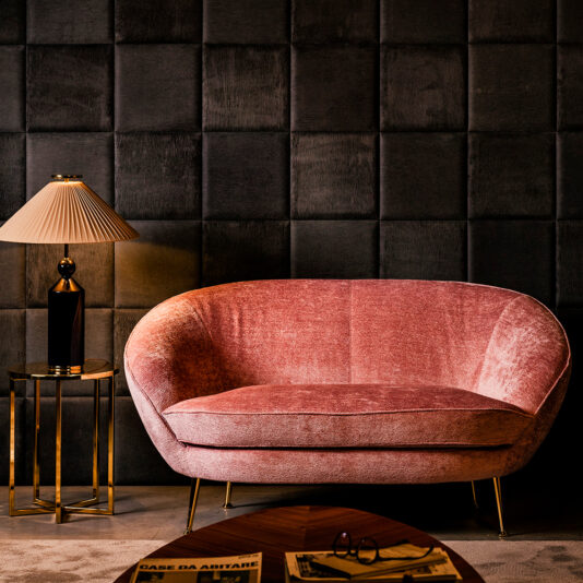 Small Contemporary Designer Sofa A cozy, stylish living room showcases the Small Contemporary Designer Sofa with plush pink velvet upholstery and gold legs. It is placed against an elegant, dark, tufted wall. To the left of the sofa is a side table adorned with a tall black and gold lamp featuring a beige pleated shade, casting a warm, ambient glow.