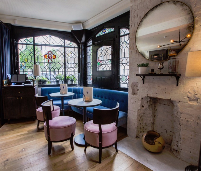 A cozy café interior with a blue velvet corner sofa, pink chairs, and small round tables. The walls feature large, decorative stained-glass windows, a white brick fireplace with a round mirror above it, and a side cabinet. Warm, inviting atmosphere.