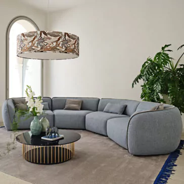 Contemporary Bouclè Modular Corner Sofa A contemporary living room showcases a large, gray Contemporary Bouclè Modular Corner Sofa with multiple cushions, accompanied by a black round coffee table with a gold geometric base adorned with decorative vases. A modern floral-patterned chandelier hangs overhead, while large leafy plants and an abundance of natural light elevate the room's ambiance.
