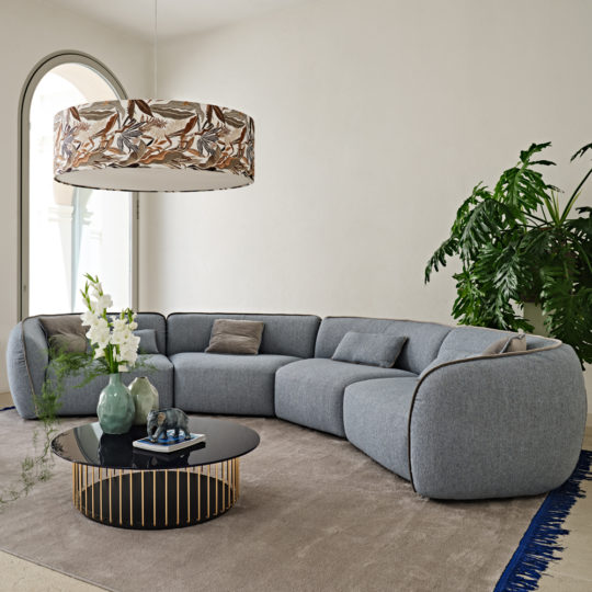 A contemporary living room showcases a large, gray Contemporary Bouclè Modular Corner Sofa with multiple cushions, accompanied by a black round coffee table with a gold geometric base adorned with decorative vases. A modern floral-patterned chandelier hangs overhead, while large leafy plants and an abundance of natural light elevate the room's ambiance.