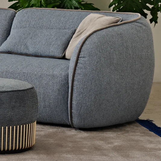 Contemporary Bouclè Modular Corner Sofa The Contemporary Bouclè Modular Corner Sofa in plush gray with rounded edges and light gray cushion sits on a beige carpet, accented by a hint of blue rug, while lush green plant leaves create a serene backdrop.