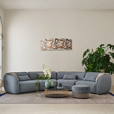 Contemporary Bouclè Modular Corner Sofa A modern living room featuring the Contemporary Bouclè Modular Corner Sofa in gray, paired with a matching round upholstered ottoman. In front of the sofa, a circular coffee table displays decorative vases and a plant. Overhead, a stylish pendant light illuminates the space, complemented by a large green plant that adorns the corner.