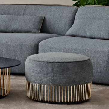Contemporary Bouclè Modular Corner Sofa A modern living room showcases a gray upholstered ottoman with a gold base, set in front of a Contemporary Bouclè Modular Corner Sofa with two cushions. A matching gold-framed coffee table peeks from the left, while a leafy plant adds freshness to the backdrop.