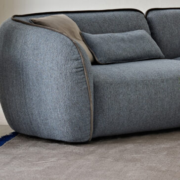Contemporary Bouclè Modular Corner Sofa Close-up of a Contemporary Bouclè Modular Corner Sofa in grey with rounded upholstery and a matching pillow. It sits on a light grey carpet with a dark blue patterned rug peeking from the corner for an elegant touch.