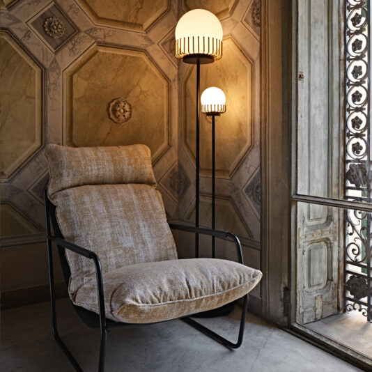 A cozy, beige upholstered armchair sits next to the Contemporary Double Sphere Floor Lamp, which features intricate line details. The background wall showcases an ornate geometric panel design, and a large window with a decorative metal grille is partially open.