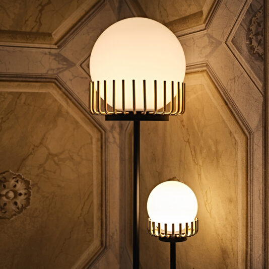 Contemporary Double Sphere Floor Lamp A warmly lit room showcases two Contemporary Double Sphere Floor Lamps, each adorned with spherical white shades and elegant gold accents. The larger lamp stands taller, while the smaller lamp is positioned just below it. Both lamps are set against an ornate, textured wall backdrop.