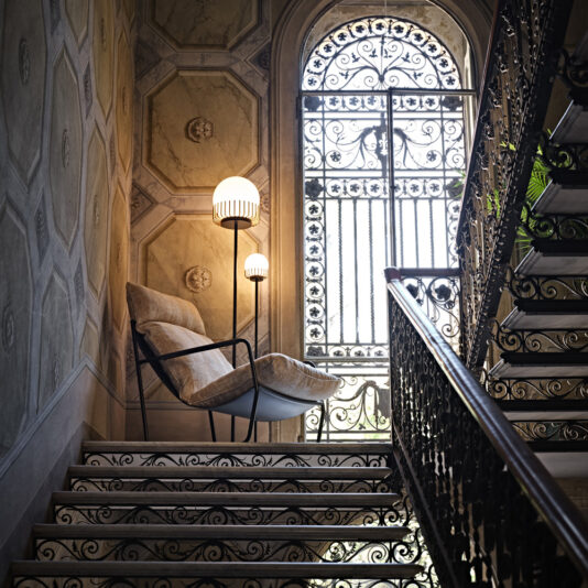 Contemporary Double Sphere Floor Lamp An elegant staircase with ornate iron railings, intricate wall designs, and a large arched window with a decorative grill. A modern chair and a Contemporary Double Sphere Floor Lamp are placed on one side of the stairway.