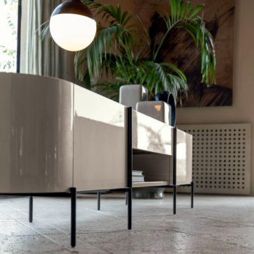 The **Contemporary Lacquered Sideboard** in glossy beige, featuring minimalist black legs, stands on a tiled floor. Potted plants and modern decor items adorn the top, while a round hanging lamp and a large window let in natural light in the background.