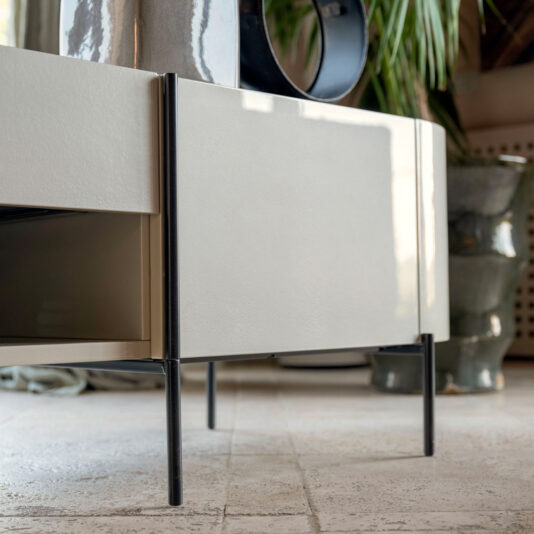 Close-up of the Contemporary Lacquered Sideboard with black legs on a tiled floor. The sideboard features a recessed space on the side, and various decorative items such as a plant and pottery are visible in the background.