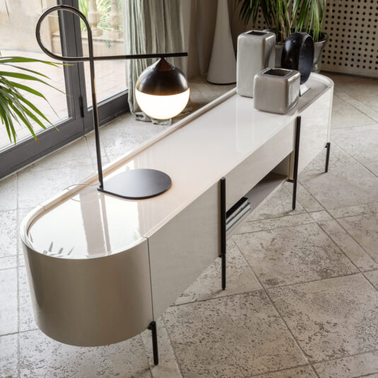 A sleek **Contemporary Lacquered Sideboard** with a glossy finish is adorned with various vases in shades of black, grey, and white. A stylish black lamp featuring a round white bulb rests on the sideboard. The setup is in a room with large floor tiles and abundant natural light.