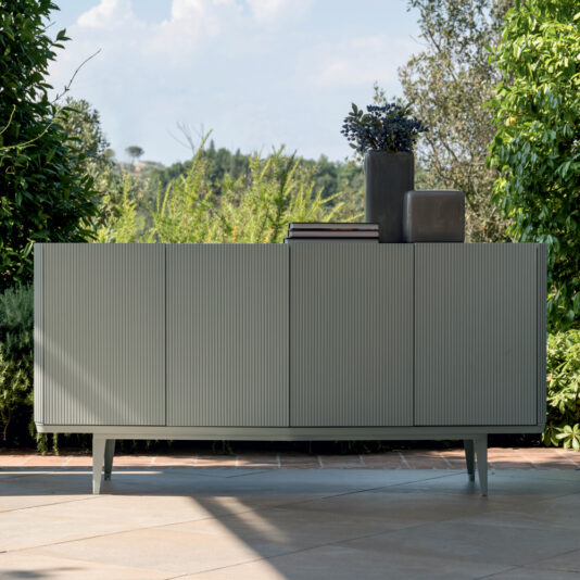 An outdoor setting featuring a Contemporary Lacquered Sideboard in a minimalist, light gray, ribbed design, standing on a tiled patio. It is adorned with two ceramic vases and a stack of books. The background includes lush greenery and a view of distant hills under a partially cloudy sky.