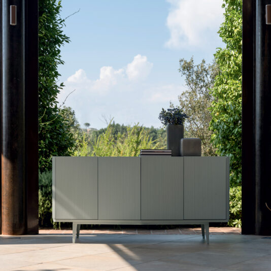 An outdoor setting showcases the Contemporary Lacquered Sideboard with vertical grooves, its light gray surface adorned with decorative vases and books. Lush green foliage and a clear blue sky are visible in the background, framed by tall, dark vertical columns.