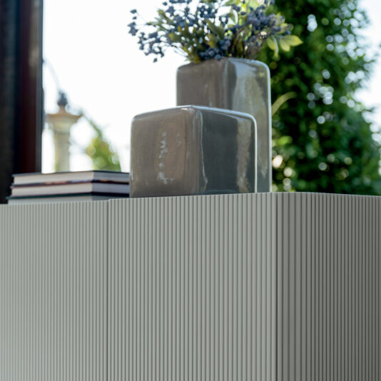 A minimalist still life showcasing a ribbed textured surface atop the Contemporary Lacquered Sideboard, adorned with a stack of books, two glossy grey ceramic vases, and a bouquet of delicate small purple flowers. The background features lush green foliage and a partial view of an outdoor structure.