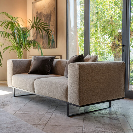 A modern living room with a textured beige sofa, two dark brown cushions, and a large glass door opening to a green, sunlit outdoor area. There's a large potted plant to the left and a piece of abstract wall art in neutral colors. The floor is tiled.