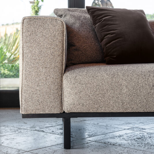 A close-up shot of a modern light grey speckled fabric couch with a plush dark brown pillow on it. The couch has black metal legs and is situated on a tiled floor. There is a large window in the background with greenery visible outside.