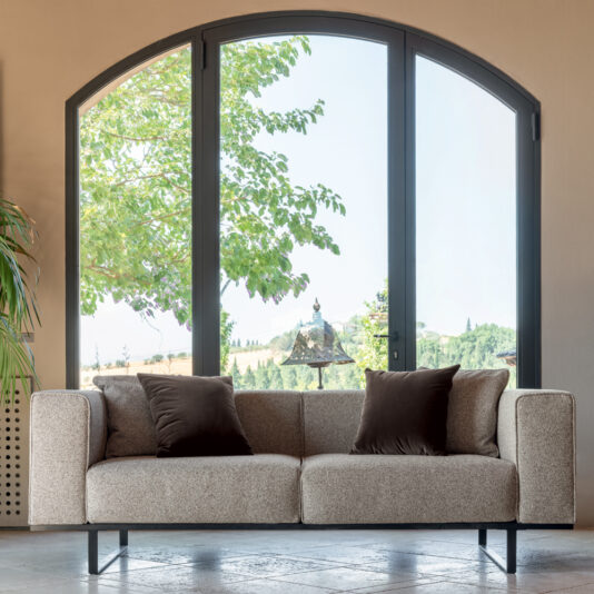 A Deluxe Fabric Upholstered Sofa in beige, adorned with dark throw pillows, is positioned in front of a grand arched window that offers views of lush greenery and rolling hills. A potted plant partially peeks into the scene from the left, adding to the cozy ambiance of the interior.