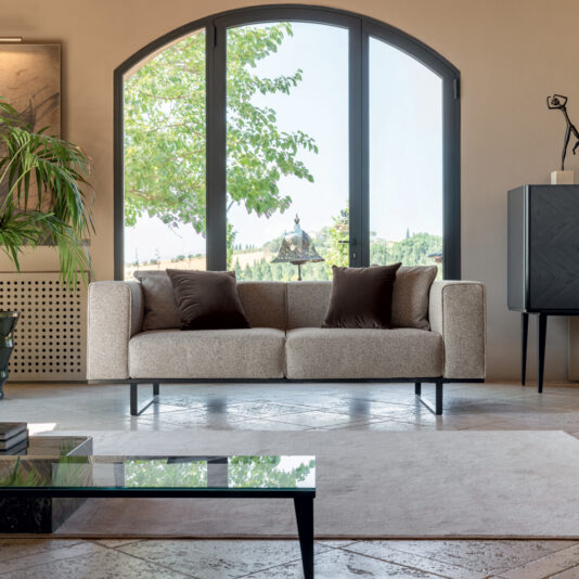A modern living room with a large arched window displaying a scenic outdoor view. The space features the Deluxe Fabric Upholstered Sofa adorned with two brown cushions, flanked by houseplants and a black cabinet. A glass coffee table sits atop a light-colored rug over a stone floor.