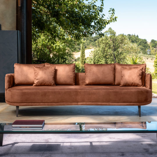 A High End Contemporary Curved Sofa in brown suede, complete with four matching cushions, is situated in a living area featuring a large open window. The serene view outside includes green trees and rolling hills under a clear blue sky. Positioned in front of the curved sofa is a transparent glass coffee table with a closed book resting on it.