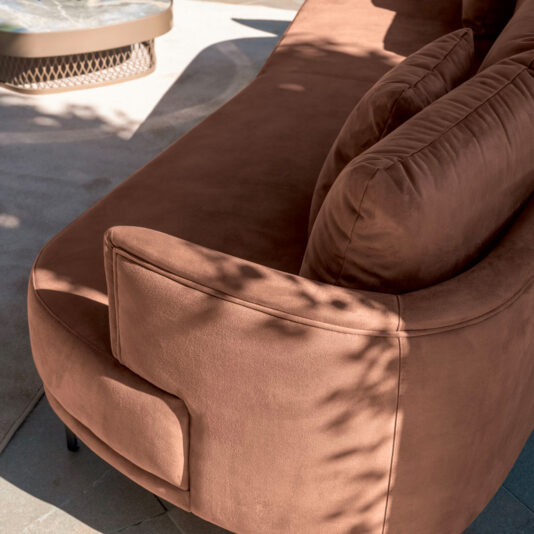 A High End Contemporary Curved Sofa, crafted from brown suede, is elegantly positioned in an outdoor setting. Sunlight filters through the leaves, casting delicate shadows on the couch. The area also features a circular coffee table with a glass top and a woven base.