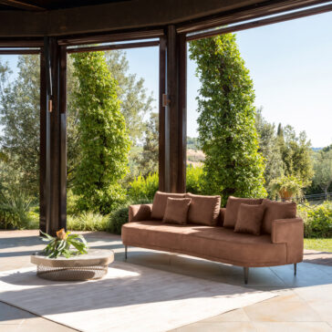 The High End Contemporary Curved Sofa with throw pillows is set on a light rug outdoors. A round coffee table with a floral centerpiece complements it, while tall trees and lush greenery stand in the background under a clear sky, enhancing the modern vibe.