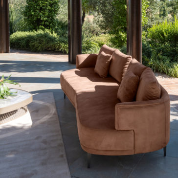 A modern outdoor seating area features the High End Contemporary Curved Sofa adorned with multiple cushions. The surrounding space is lush with greenery, including well-manicured shrubs and trees, creating a serene garden setting. A round white table is set to the side on a beige rug.