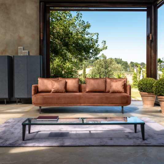 A high-end contemporary living room with an open wall features a High End Contemporary Curved Sofa, a glass coffee table on a lavender rug, and a view of lush greenery outside. Potted plants and grey storage units against the wall seamlessly blend indoor comfort with outdoor scenery.