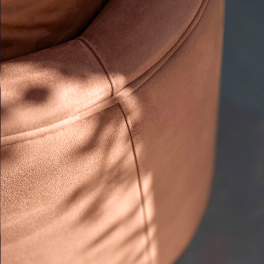 Close-up of the High End Contemporary Curved Sofa, showcasing its soft, light brown color illuminated by sunlight. Shadow patterns fall across the surface, creating diagonal lines and subtle shading. The light highlights the fabric's smooth texture and detailed seam.