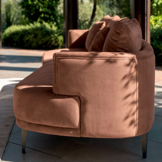 A High End Contemporary Curved Sofa upholstered in luxurious brown suede is perfectly placed outdoors beneath shaded wooden beams. Plush cushions beckon for relaxation as sunlight filters through, casting gentle shadows. The serene backdrop of lush green plants and blurred trees completes this inviting setting.