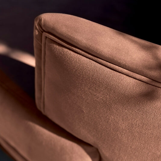 Close-up shot of the backrest of a pink, velvety chair. The fabric appears smooth and slightly shiny, reminiscent of the High End Contemporary Curved Sofa, with soft shadows and light creating a warm ambiance.