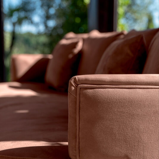 Close-up of a corner of the High End Contemporary Curved Sofa in a well-lit room. The plush brown sofa is adorned with matching brown cushions. A large window in the background offers a blurred view of green foliage, indicating an outdoor garden scene.