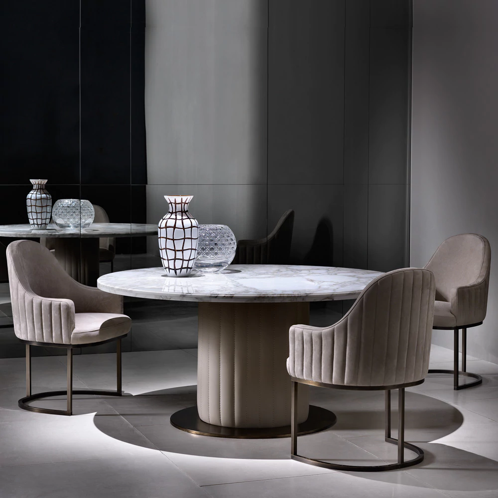 A modern, elegant room featuring a round marble table with a central column base, surrounded by three beige upholstered chairs with metal frames. On the table are two decorative vases. The room has mirrored walls, reflecting the table and chairs.
