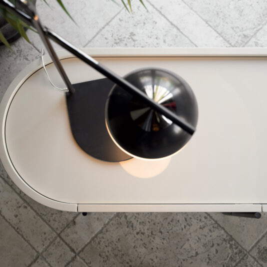 A Luxury Iron And Brass Table Lamp with a round shade is illuminated on an oval-shaped white desk. The lamp casts a warm glow on the desk's surface. The photo is taken from an overhead perspective, and part of a plant is visible on the left side.