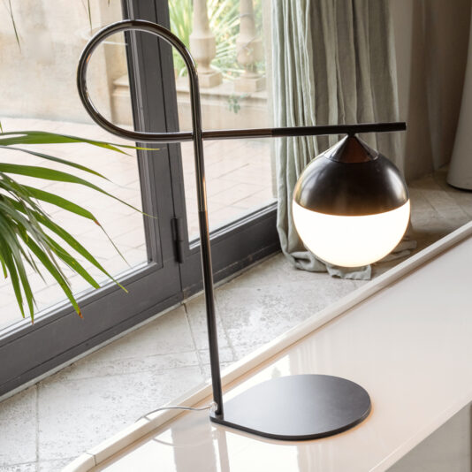 The Luxury Iron And Brass Table Lamp boasts a sleek, modern design with a black metallic frame. Its elegance is highlighted by its circular base and horizontal arm, which supports a spherical light featuring a two-tone design: the top half is black and the bottom half is white. Positioned on a white surface near a window, this lamp exudes sophistication and style.