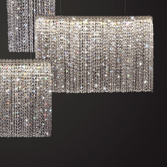 A close-up showcases three Luxury Narrow Cut Crystal Chandeliers, each emitting a sparkling glow against a dark background. These exquisite chandeliers are adorned with numerous hanging crystal strands that beautifully reflect light, creating a dazzling display. The overall aesthetic is both elegant and glamorous.