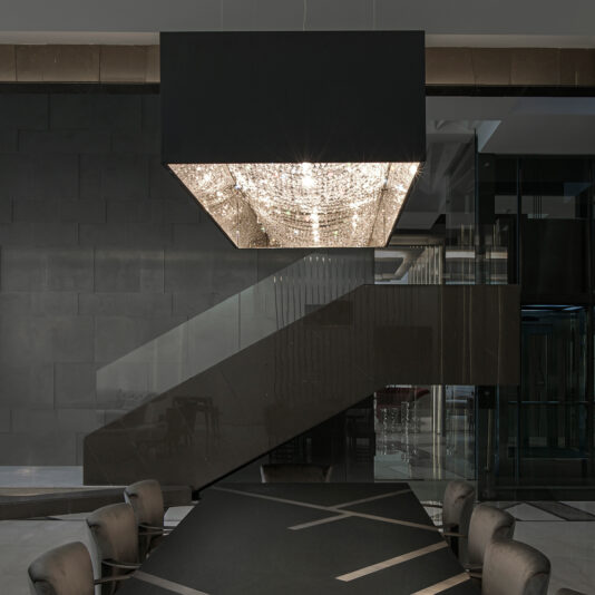 Luxury Rectangular Crystal Chandelier A modern, minimalistic dining area with a dazzling Luxury Rectangular Crystal Chandelier hanging above the black dining table. The room features a sleek, black staircase against a dark wall, creating a sophisticated ambiance.